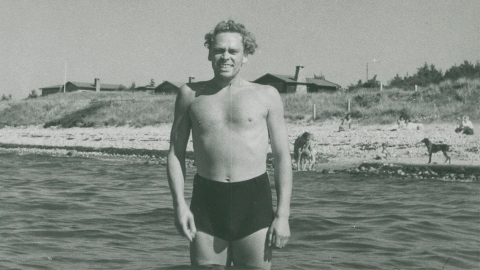 En ung Dirch Passer i badebukser står i vandet foran en strand. 